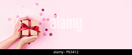 Woman's Hände, die Kraft Geschenkbox mit roter Schleife auf rosa Hintergrund mit Konfetti eingerichtet.. Ansicht von oben, Urlaub vorliegenden Konzept mit Kopie Raum, Banner. Stockfoto