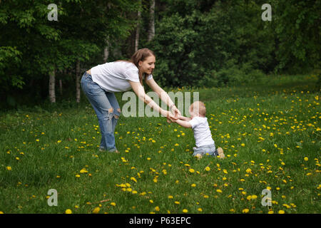 Familie. Mutter und Sohn gehen herum Wiese halten sich an den Händen. Schwangere Mädchen hilft kleinen Jungen laufen lernen. Gelber Löwenzahn wächst in der Wiese. Stockfoto