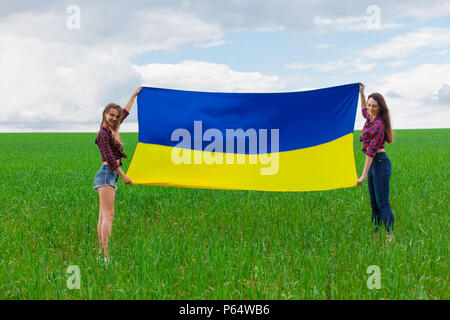 Zwei junge schöne ukrainische Mädchen mit einem ausgezeichneten Abbildung entfaltete die Ukrainische gelbe und blaue Flagge gegen den blauen Himmel und die grüne g Weizen fie Stockfoto