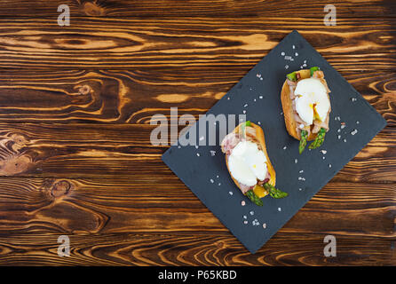 Gegrillte Toast mit Spargel in Schinken und pochiertem Ei auf hölzernen Hintergrund Stockfoto