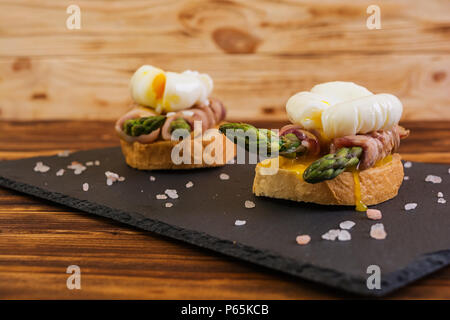 Gegrillte Toast mit Spargel in Schinken und pochiertem Ei auf hölzernen Hintergrund Stockfoto