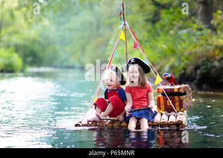 Kinder gekleidet in Piraten Kostüme und Hüte mit Schatztruhe, Ferngläser und Schwerter spielen auf hölzerne Floß Segeln in einem Fluss an heißen Sommertag. Pira Stockfoto