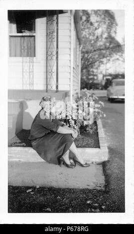 Schwarz-weiß Foto, zeigt eine attraktive, lächelnde Frau mit kurzen, lockige, blonde Haare, trug einen Rock und Highheels, und sitzen auf die konkrete Grenze ein Blumenbeet vor einem kleinen Haus, mit einem Oldtimer im Hintergrund sichtbar, wahrscheinlich in Ohio in den zehn Jahren fotografierte nach dem Zweiten Weltkrieg, 1950. () Stockfoto