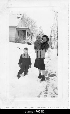 Schwarz-weiß Foto, zeigt eine dunkelhaarige Frau, in voller Länge, trägt eine Baskenmütze, kurze Stiefel, Rock und Jacke, stehend auf einem verschneiten Gehweg, mit einer kleinen, gemütlich Kind in ihren Armen gekleidet, mit einem zweiten Junge, trägt ein Schneeanzug, Mütze, stand neben ihr, mit schneebedeckten Bäumen und einem Abstellgleis bedeckt im Hintergrund, wahrscheinlich in Ohio in den zehn Jahren fotografierte nach dem Zweiten Weltkrieg, 1950. () Stockfoto