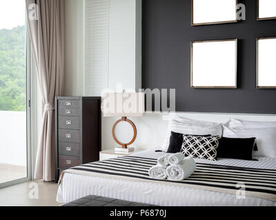 Modernes Schlafzimmer mit vier Quadratmeter leer Edelstahl Rahmen auf die graue Wand Innenraum, Kopfkissen und Handtücher auf dem Bett mit seitlichen Leuchte und Holz- vintag Stockfoto