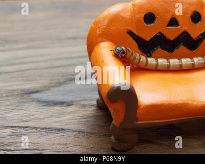 Super oder riesigen Wurm kriecht auf orange Miniatur Keramik Kürbis Couch oder Sofa über dunklen Holz- Oberfläche mit Kopie Raum als Hintergrund in Halloween, Ornament, Feier, und Dekoration Stockfoto