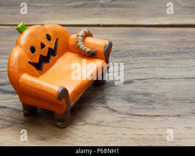 Super oder riesigen Wurm kriecht auf orange Miniatur Keramik Kürbis Couch oder Sofa über dunklen Holz- Oberfläche mit Kopie Raum als Hintergrund in Halloween, Ornament, Feier, und Dekoration Stockfoto