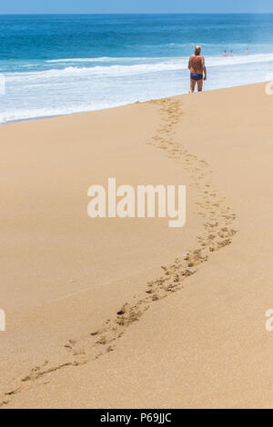 Eine tropische Szene als ein älterer Mann auf einem Strand hinterlässt Spuren im Sand mit einem strahlend blauen Meer und weißen Wellen schäumen im Hintergrund. Stockfoto