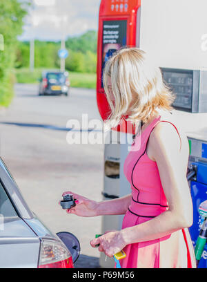 Weiß, attraktive kaukasisch Weiblich 29 Jahre alt mit blonden Haaren, in rosa Kleid, schließen Auto Tank mit Stecker nach dem Tanken an der Tankstelle. Stockfoto