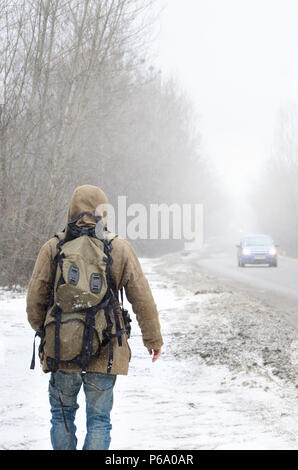 Ein Mann mit einem großen Rucksack Spaziergänge entlang eine asphaltierte Straße im Winter bei einem Blizzard. Stockfoto