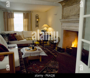 Weiße Sofas und einem Couchtisch mit einem weißen Tuch und eine Flasche Wein im Cottage Wohnzimmer mit brennenden Feuer im Kamin aus Stein Stockfoto