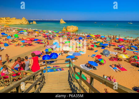 Lagos, Portugal - 21 August 2017: holzsteg Fußgängerbrücke zum malerischen Strand Praia Dona Ana. Menschen Sonnenbaden und genießen unter bunten Sonnenschirmen in beliebten Strand Dona Ana. Sommer Urlaub an der Algarve. Stockfoto