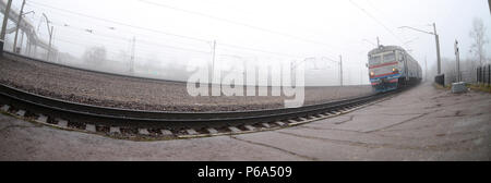 Die Ukrainische s-bahn eilt entlang der Bahnstrecke in einem nebligen Morgen. Fisheye Foto mit erhöhter Verzerrung. Stockfoto