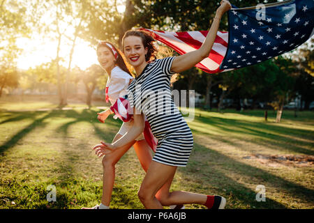 Schöne junge Frau mit Freundin, die in den Park amerikanische Flagge. Zwei Mädchen mit USA-Flagge im Freien genießen. Stockfoto