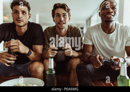 Freunde Spaß mit Getränken und Snacks beim Spielen von Spiel zu Hause. Zwei Männer spielen video game Holding Joysticks, während ein anderer Mann Uhren Esprit Stockfoto