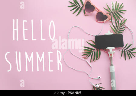 Hallo Sommer text auf stilvolle rosa Sonnenbrille, Telefon auf selfie Stick, Kopfhörer, und grüne Palmen auf rosa Hintergrund. Stilvolle Sommerferien fla Stockfoto