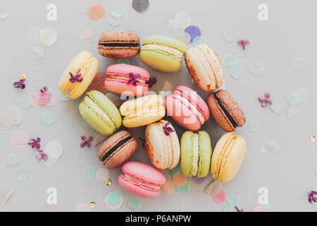 Lecker Pink, Gelb, Grün und Braun makronen am trendigen Pastellfarben grauem Papier mit lila Blumen und Konfetti. Platz für Text. leckere bunte Makronen Stockfoto