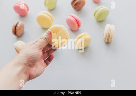 Hand mit köstlichen gelben Makronen mit Rissen am trendigen Pastellfarben grau Papier. Lecker Pink, Gelb, Grün und Braun Makronen im Hintergrund. Platz für Stockfoto