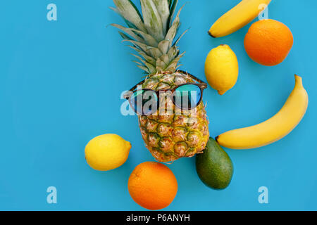 Ananas in modischer Sonnenbrille, und Bananen, Orangen-, Avocado- und Zitronen auf hellen blauen Papier, trendige Flach. Früchte modernes Image, Top View. saftig Stockfoto