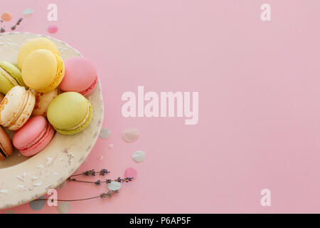 Leckere bunte Macarons in vintage Platte am trendigen Pastellfarben rosa Papier mit Lavendel und Konfetti. Platz für Text rosa, gelb, grün, weiß, braun Maca Stockfoto