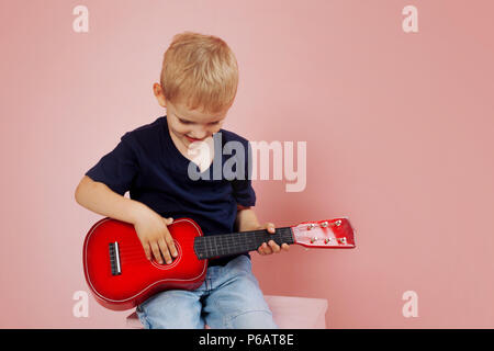 Kleiner Junge lernt auf einer kleinen Gitarre zu spielen. Studie Musik. Ukulele. Portrait auf rosa Hintergrund Stockfoto