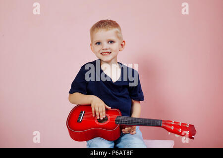 Kleiner Junge lernt auf einer kleinen Gitarre zu spielen. Studie Musik. Ukulele. Portrait auf rosa Hintergrund Stockfoto