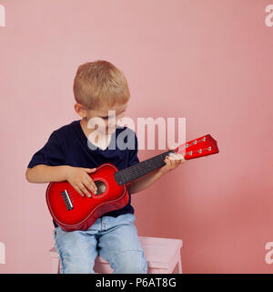 Kleiner Junge lernt auf einer kleinen Gitarre zu spielen. Studie Musik. Ukulele. Portrait auf rosa Hintergrund Stockfoto