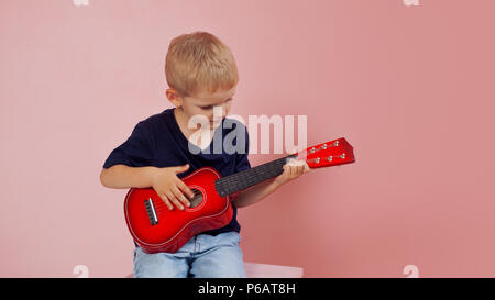 Kleiner Junge lernt auf einer kleinen Gitarre zu spielen. Studie Musik. Ukulele. Portrait auf rosa Hintergrund Stockfoto