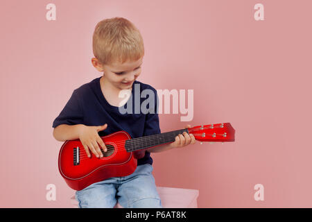 Kleiner Junge lernt auf einer kleinen Gitarre zu spielen. Studie Musik. Ukulele. Portrait auf rosa Hintergrund Stockfoto