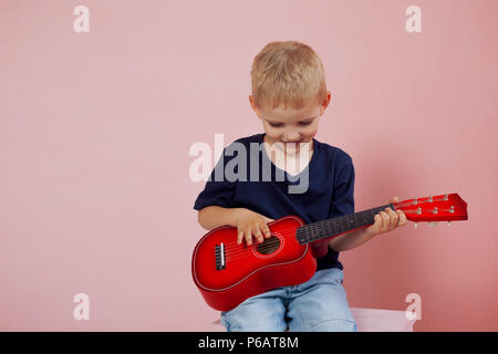 Kleiner Junge lernt auf einer kleinen Gitarre zu spielen. Studie Musik. Ukulele. Portrait auf rosa Hintergrund Stockfoto