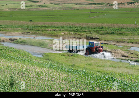 Traktor fahren durch ein bewässerungskanal, Ziehen einer Last, die eine Welle und Spritzwasser vorne, in Nordkorea DIE DEMOKRATISCHE VOLKSREPUBLIK KOREA Stockfoto