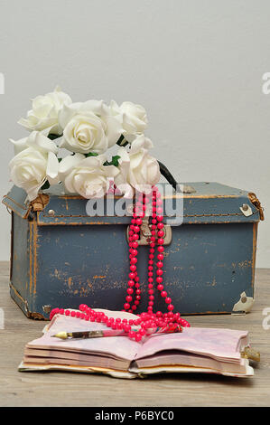Eine alte abgenutzte blaue Koffer mit einer Reihe von künstlichen weissen Rosen, ein String rosa Perlen, ein Notizbuch und einen Stift Stockfoto