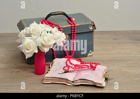 Eine alte abgenutzte blaue Koffer mit einer Reihe von künstlichen weissen Rosen, ein String rosa Perlen, ein Notizbuch und einen Stift Stockfoto