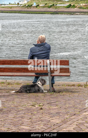 Mann sitzt auf einer Holzbank mit seinem Hund, der auf das Meer Stockfoto