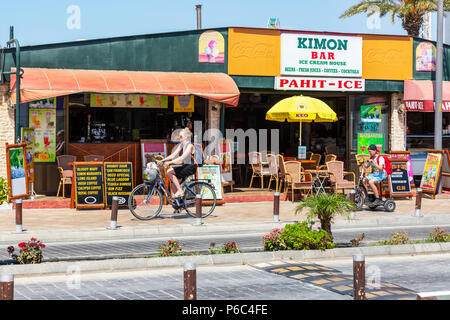 Frau auf einem Fahrrad von ihrem Sohn auf einem elektrischen Trike gefolgt, Radfahren auf einem Radweg vor einem öffentlichen Bar, Ayia Napa, Zypern Stockfoto