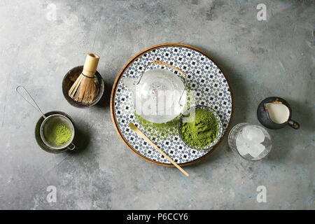 Zutaten für die Herstellung von Matcha Eis trinken. Grüner Tee matcha Pulver in Keramik Schüssel, traditionelle Bambus Löffel, Schneebesen auf Platte, Teekanne aus Glas, Eiswürfel ov Stockfoto