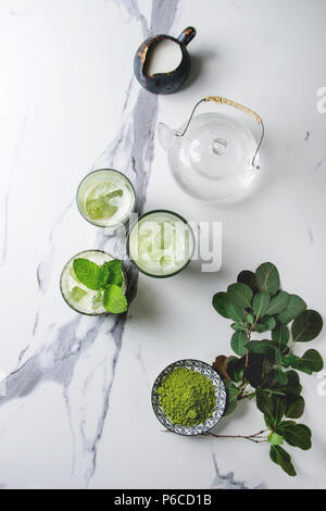 Matcha Grüntee Iced Latte oder Cocktail in drei verschiedene Gläser mit Eiswürfeln, Matcha Pulver und kanne Milch auf weißem Marmortisch, durch g eingerichtet Stockfoto