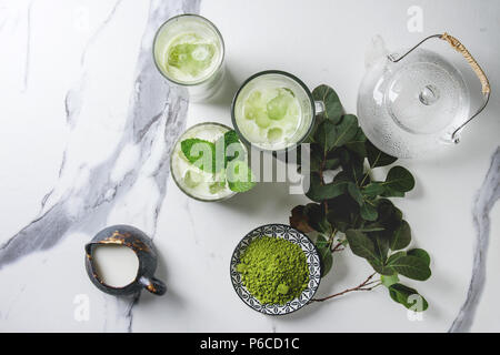 Matcha Grüntee Iced Latte oder Cocktail in drei verschiedene Gläser mit Eiswürfeln, Matcha Pulver und kanne Milch auf weißem Marmortisch, durch g eingerichtet Stockfoto