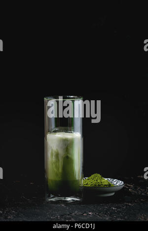 Matcha Grüntee Iced Latte oder Cocktail in hohes Glas mit Eiswürfeln und Matcha Pulver über dunkle Textur Hintergrund Stockfoto