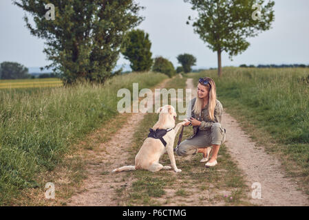 Liebevolle junge Frau eine Pfote von ihrem Hund angeboten als Sie kauert auf einem Feldweg in der Landschaft bei Ihren täglichen Spaziergang Stockfoto