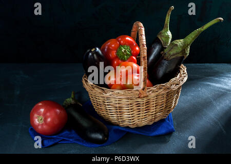 Gemüse in Weidenkorb und Auberginen und Tomaten auf blau serviette auf dunklem Hintergrund. Stockfoto