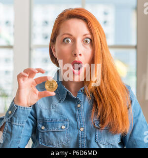Rothaarige Frau mit bitcoin cryptocurrency zu Hause Angst im Schlag mit einer Überraschung konfrontiert, ängstlich und aufgeregt mit Angst Ausdruck Stockfoto