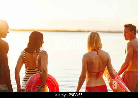 Rearview eine vielfältige Gruppe von jungen Freunden in Badebekleidung gemeinsam in einem See den Sonnenuntergang zu beobachten. Stockfoto