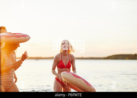 Lachende junge blonde Frau in einem Bikini, in einem See mit einem Ring schwimmen und dabei Spaß haben mit Freunden an einem See bei Sonnenuntergang Stockfoto