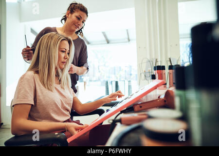 Blonde junge Frau sitzt in einem Salon Stuhl durch Haarfarbe Proben in einem Buch mit ihr Hairstylist Stockfoto