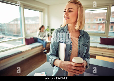 Lächelnden jungen Geschäftsfrau mit einem Kaffee und, einen Laptop, während in einem Büro mit Kollegen im Hintergrund Stockfoto