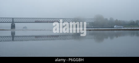 Nebligen Herbstmorgen in Deutschland am Rhein, eine fließende Barge in der Ferne eine alte Eisenbahnbrücke. Stockfoto