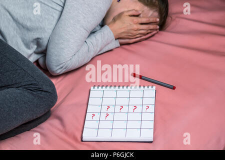 Der Kalender Zeitraum mit markierten Fragezeichen liegt in der Nähe der jungen traurige Frau. Ein depressiver Mädchen, eine ungeplante Schwangerschaft. Verzögerung der Menstruation. Stockfoto