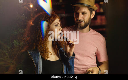 Liebespaar auf Datum. Kerl und Freundin Rauch elektronische Zigaretten. Sie sprechen. Rothaarige Mädchen. Mann in Hut und T-Shirt. Lichter der Nacht Stadt Stockfoto