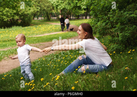 Junge schwangere Mädchen sitzt auf Rasen im City Park. Sohn hält die Hand seiner Mutter. Er ruft Sie mit ihm spazieren zu gehen. Stockfoto